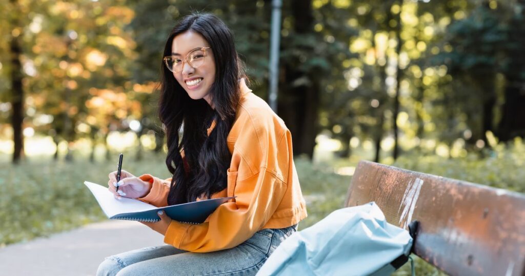 Young woman with glasses smiling while writing in a notebook on a park bench.